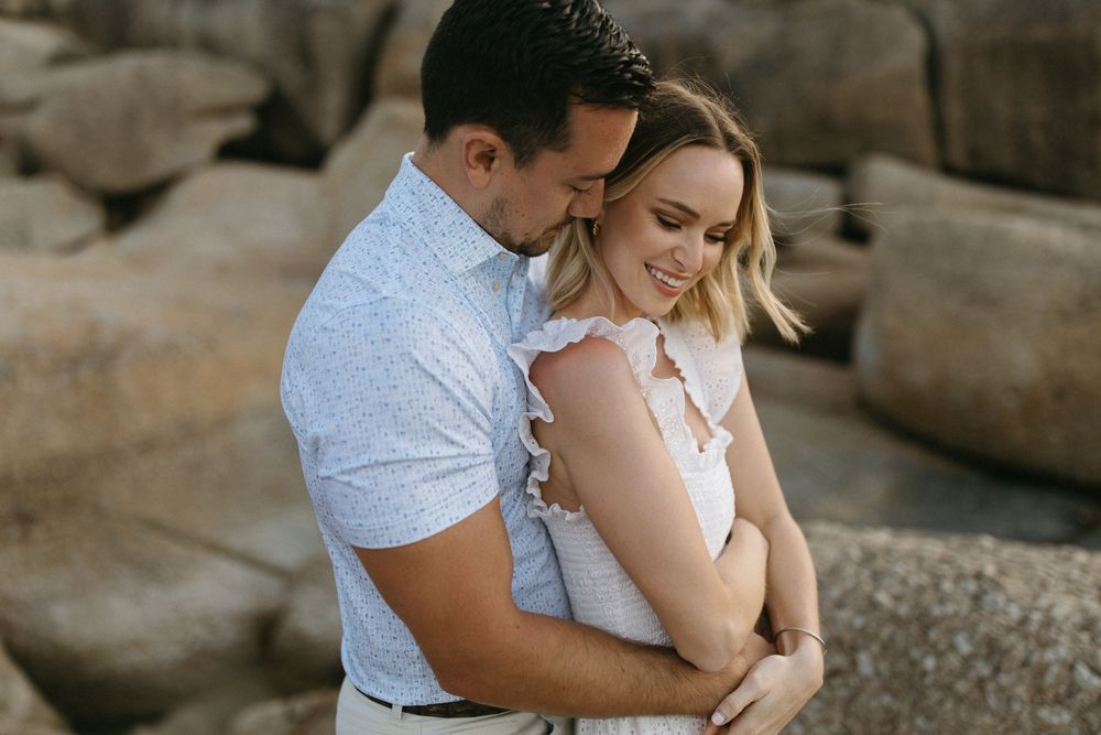 Couple embracing romantically outdoors near rocky background, man kissing woman's head while she smiles softly.