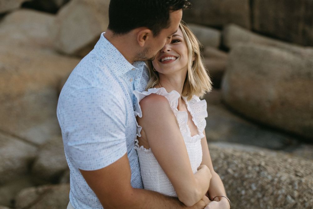 A smiling couple embraces romantically outdoors near rocky terrain, sharing a tender moment together.