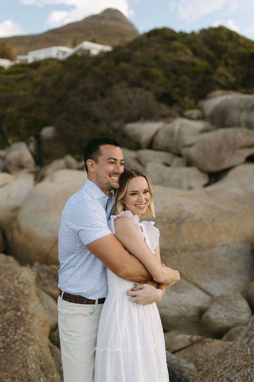 A smiling couple embraces on a rocky coastal landscape, the woman in white dress and man in light blue shirt, with mountains behind.