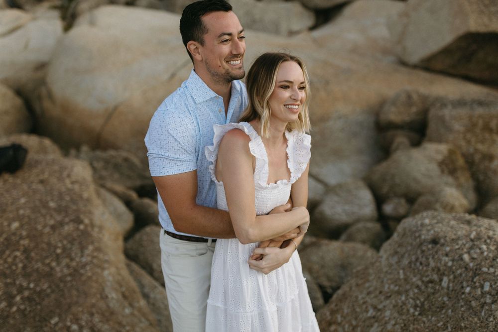 Couple embracing on rocky beach, woman in white dress smiling, man in light blue shirt, outdoor engagement photo.