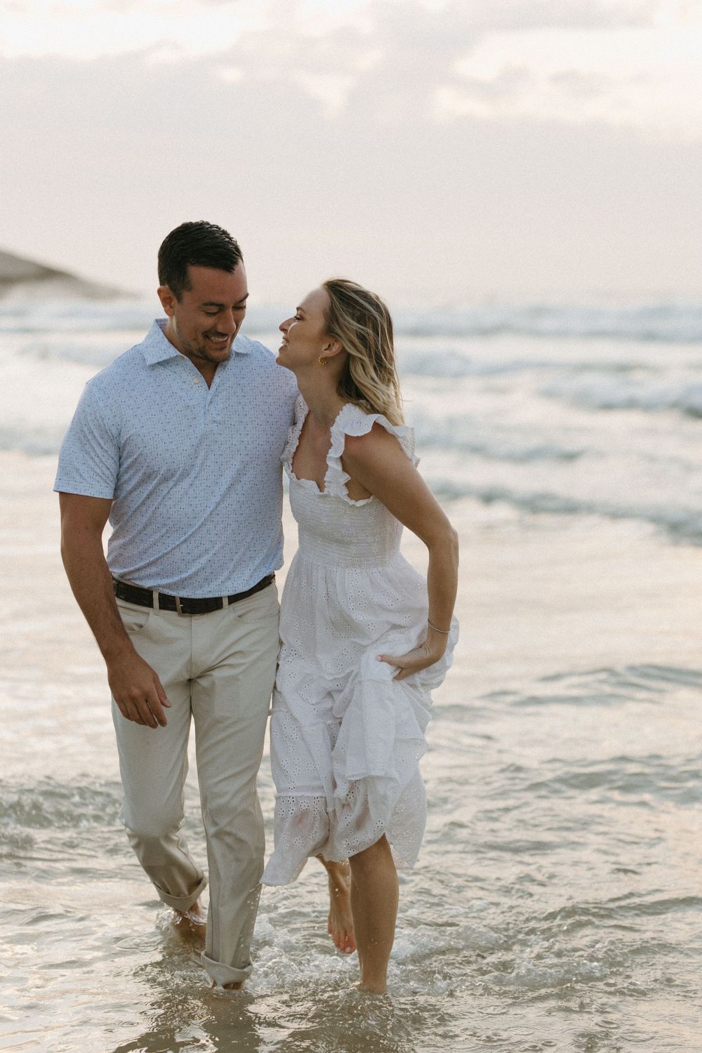 Couple laughing and walking barefoot together in ocean waves at sunset beach.