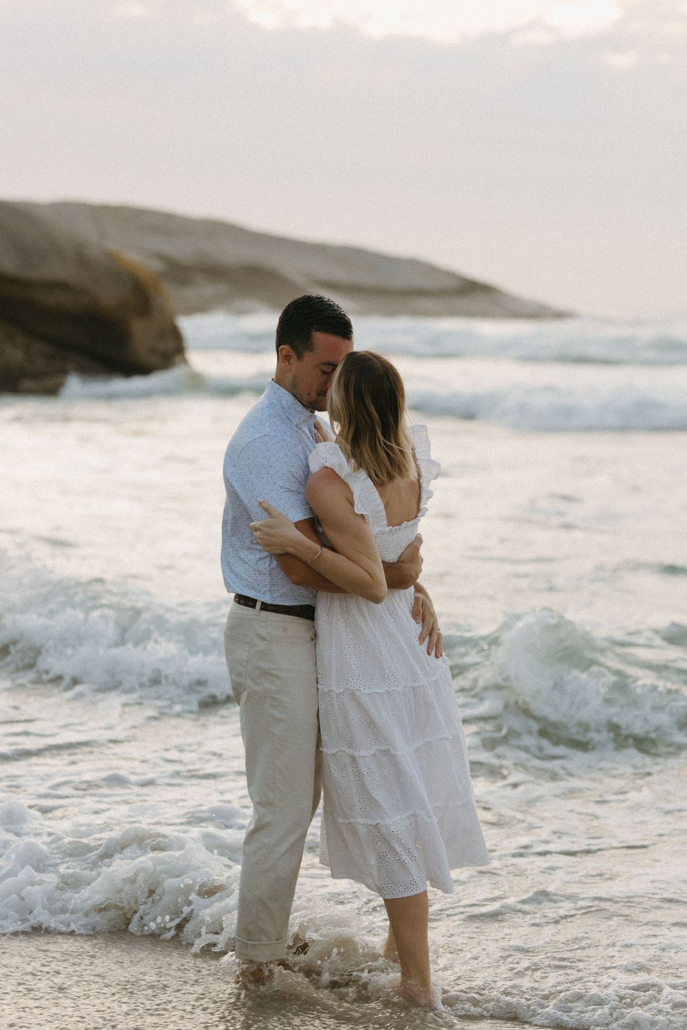 Romantic couple embracing on a beach shore with ocean waves, man in blue shirt and woman in white dress.