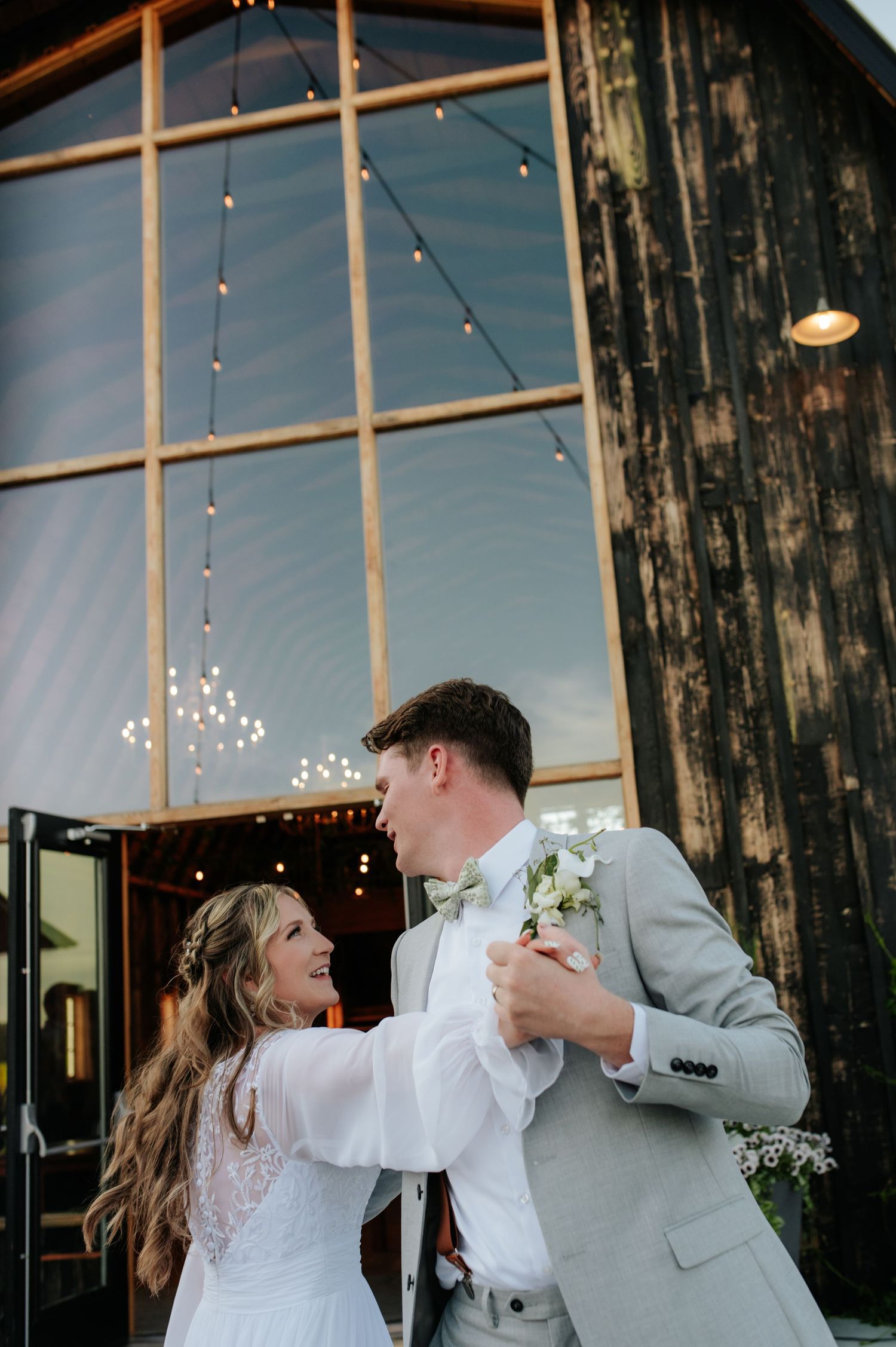 Bride and groom share romantic embrace in rustic barn venue with wood beam ceiling and string lights.