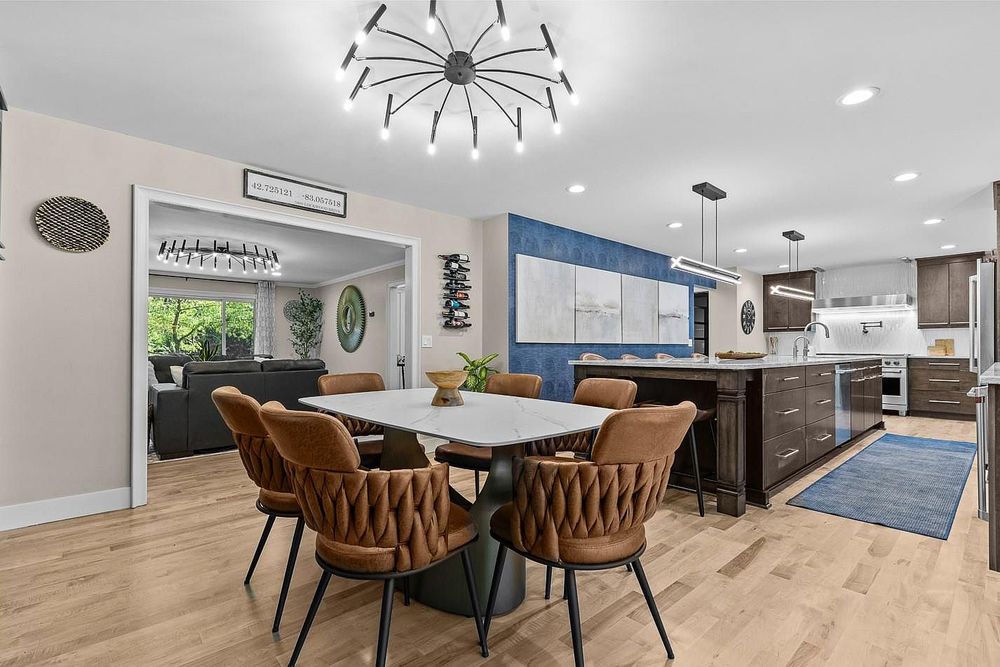 Modern open-concept dining area with brown velvet chairs, white table, chandelier, and kitchen with blue tile backsplash.