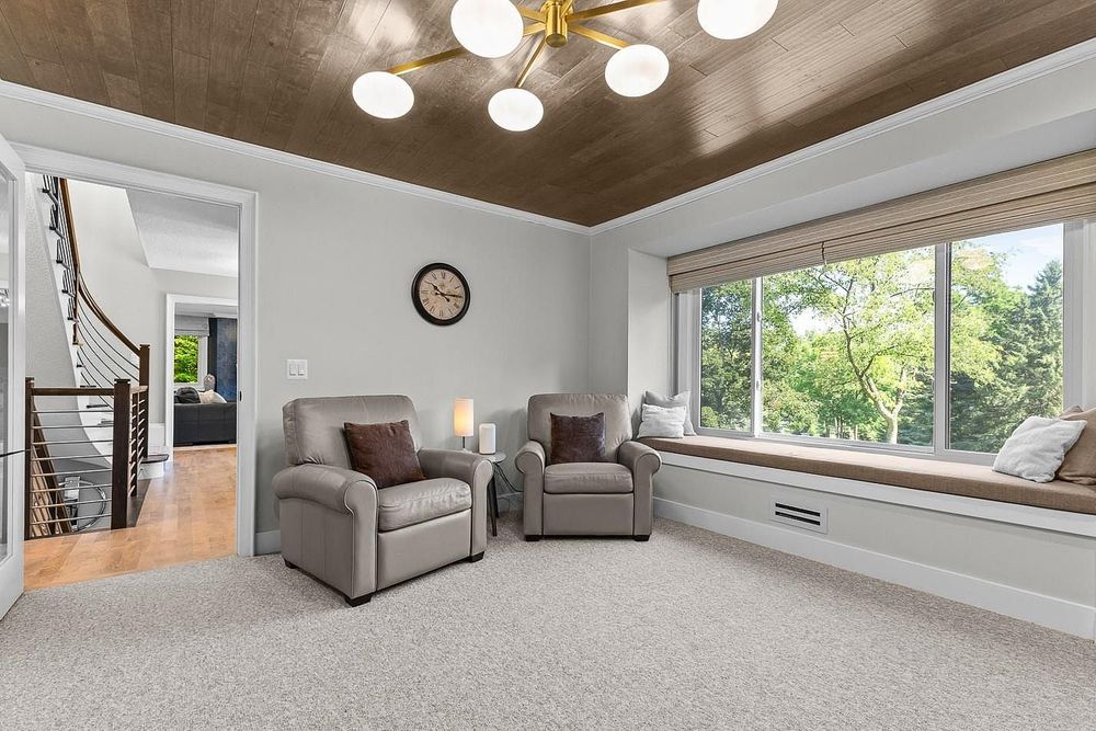 Cozy living room with gray armchairs, built-in window seat, wood tray ceiling, and large windows overlooking lush greenery.
