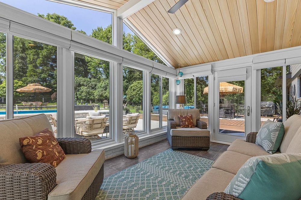 Bright sunroom with wicker furniture, vaulted wood ceiling, large windows, and view of outdoor pool and patio.