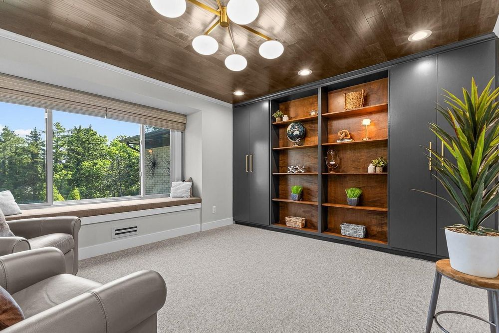 Modern home office with built-in dark shelving, wood ceiling, large window seat, and plush gray carpet flooring.