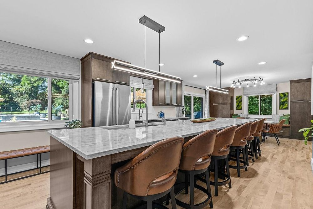 Modern kitchen with large marble island, brown leather bar stools, dark wood cabinets, and stainless steel appliances.