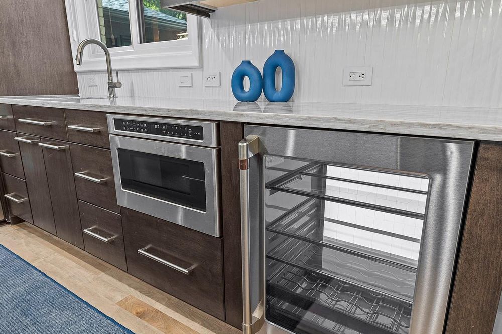 Modern kitchen with dark wood cabinets, stainless steel oven, open wine fridge, and white quartz countertops.