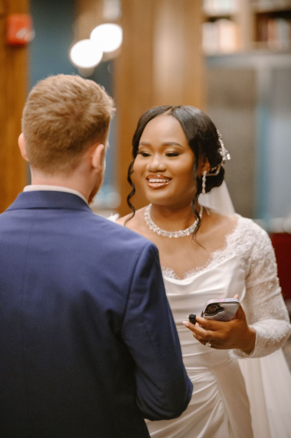 Smiling bride in lace wedding gown holding vows card facing groom in blue suit during ceremony.