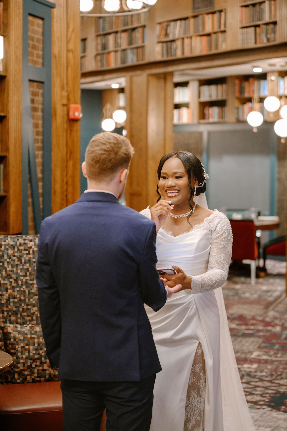 Smiling bride in lace gown shares a joyful moment with groom in navy suit in library setting.