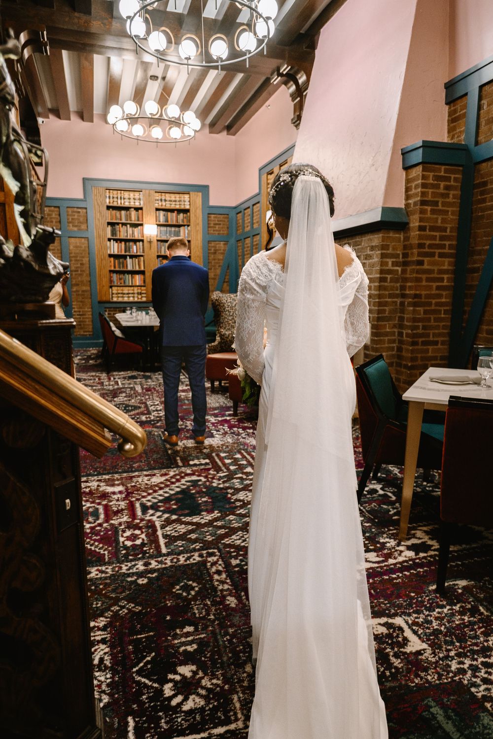 Bride in white gown and veil stands in elegant library as groom in navy suit waits ahead.