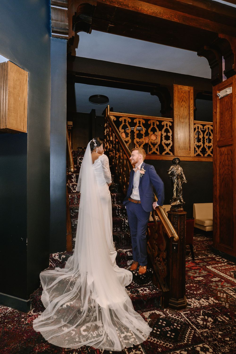 Bride in white gown with veil and groom in navy suit share a moment at ornate wooden staircase.