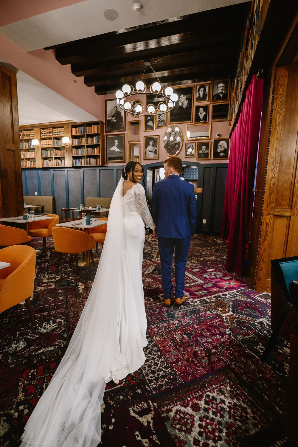 Bride and groom holding hands walking through an elegant library venue with ornate rugs and portrait gallery walls.