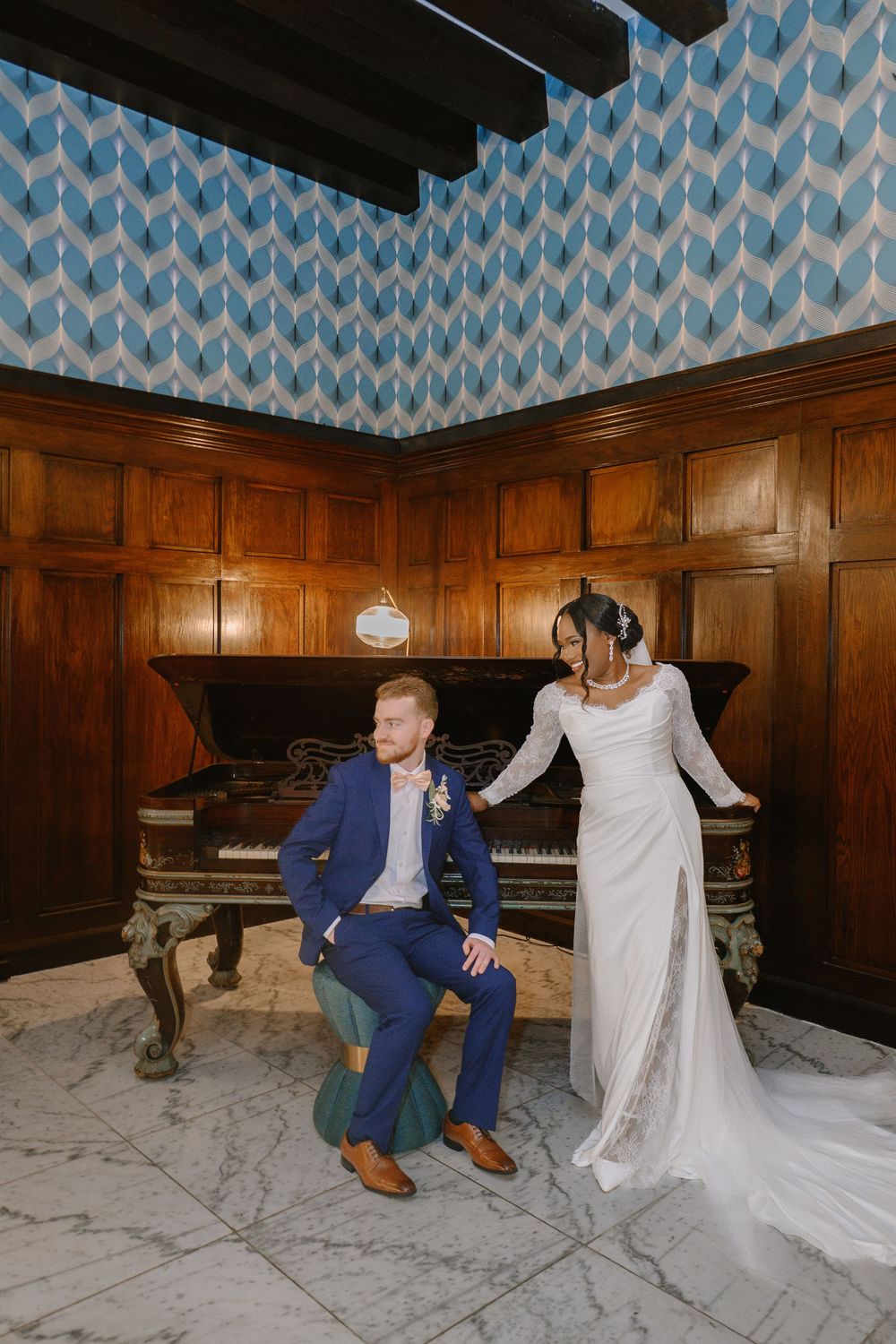 Bride and groom pose by grand piano in wood-paneled room with blue geometric ceiling.