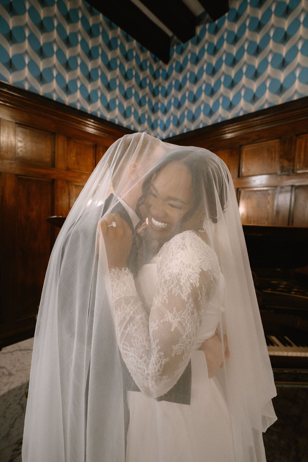 Joyful bride and groom share an intimate moment under a white wedding veil, laughing together in an elegant wood-paneled room.