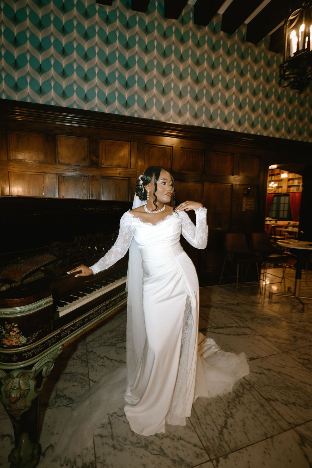 Elegant bride in long-sleeve white gown with train poses beside a grand piano in a wood-paneled ballroom.