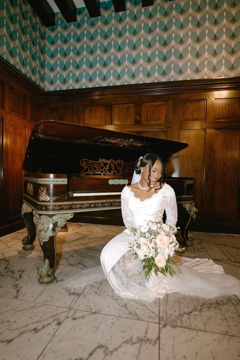 Bride in white gown sits on marble floor beside ornate grand piano with floral bouquet.