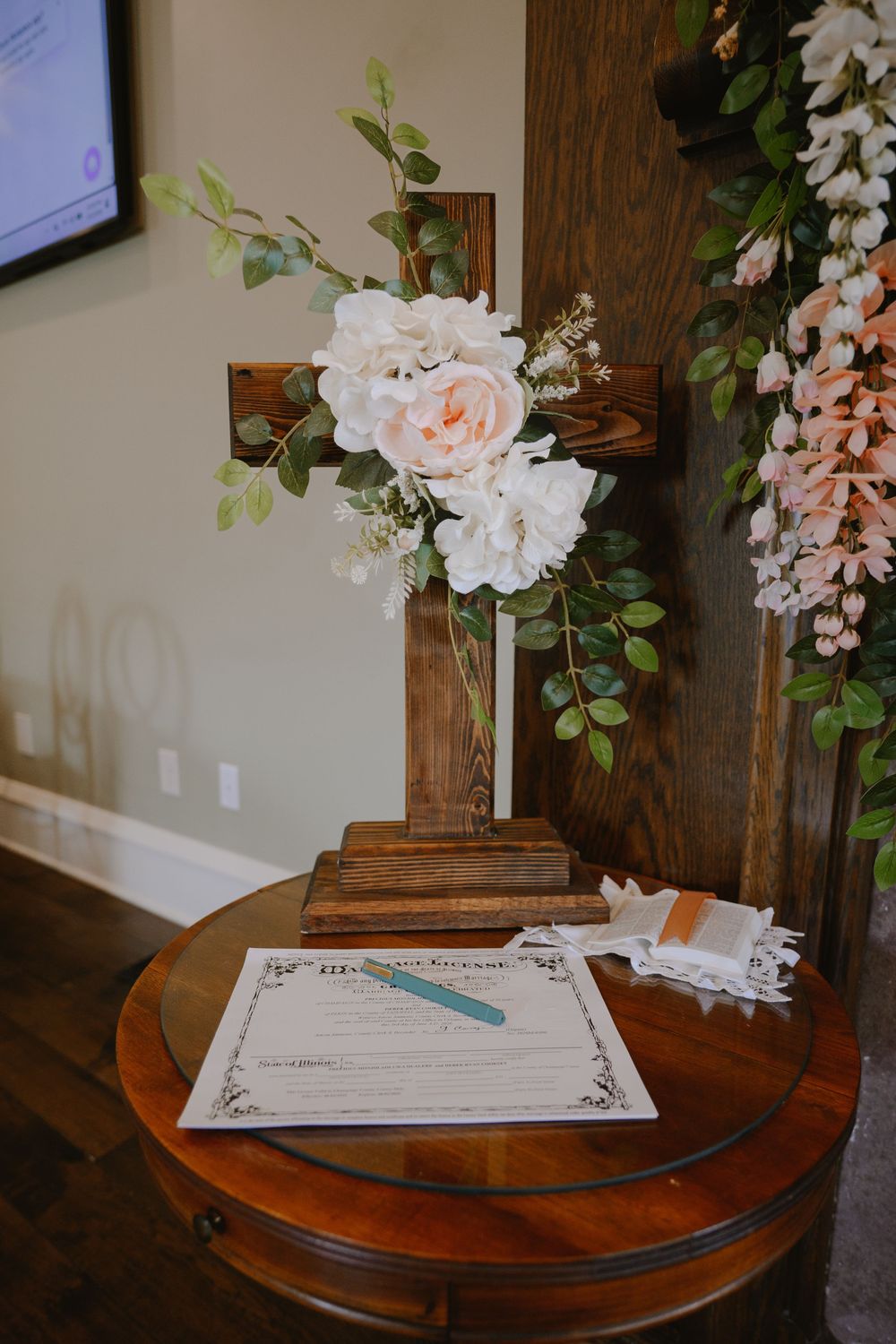 Wooden cross with white floral arrangement beside a marriage license on a round table at a wedding venue.
