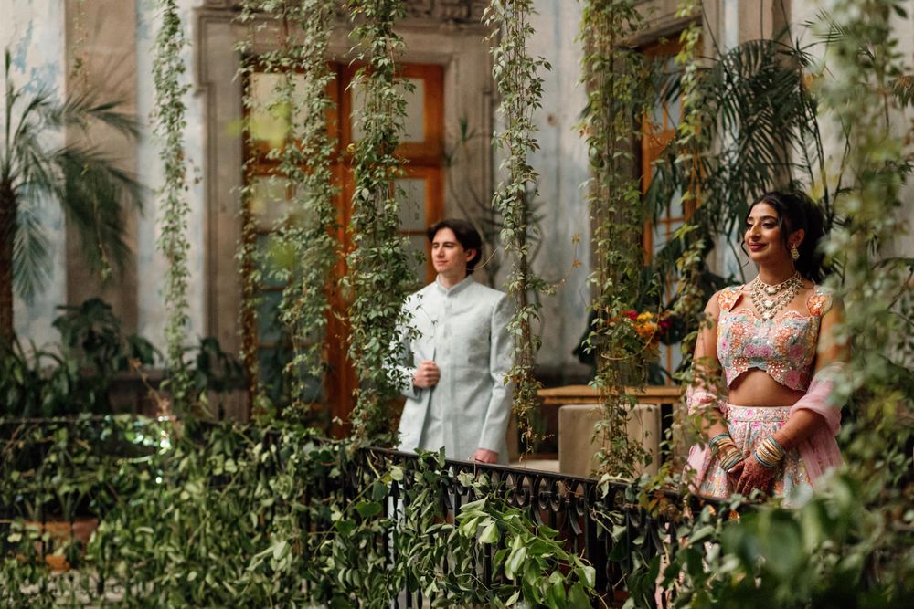 Couple in traditional South Asian attire standing on a lush green balcony adorned with climbing vines.