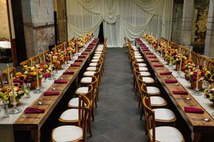 Elegant banquet hall with long wooden tables, white chairs, burgundy napkins, and vibrant floral centerpieces in red and yellow.