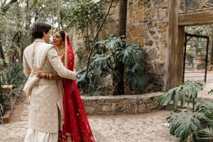 Couple in traditional Indian wedding attire embracing outdoors, bride in red saree, groom in cream sherwani.