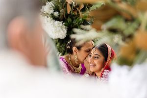 Two women in vibrant Indian bridal attire share a joyful moment during a traditional wedding ceremony.
