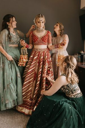 Bride in red lehenga with gold jewelry surrounded by bridesmaids in green and grey outfits during Indian wedding preparation.