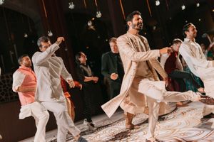 Men in traditional South Asian attire perform energetic dance moves at a festive wedding celebration.