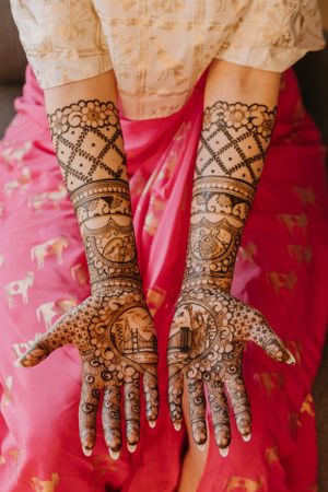 Intricate henna designs cover both hands and forearms against pink fabric background.