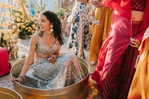 Bride in silver lehenga sits in traditional metal basin during wedding ceremony.