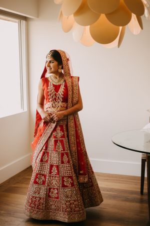 A bride in a traditional red and gold embroidered lehenga poses elegantly in a minimalist room with wooden floors.