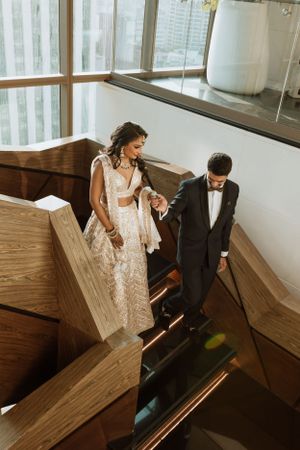 Couple in formal wedding attire descending wooden stairs in a modern building with large windows.