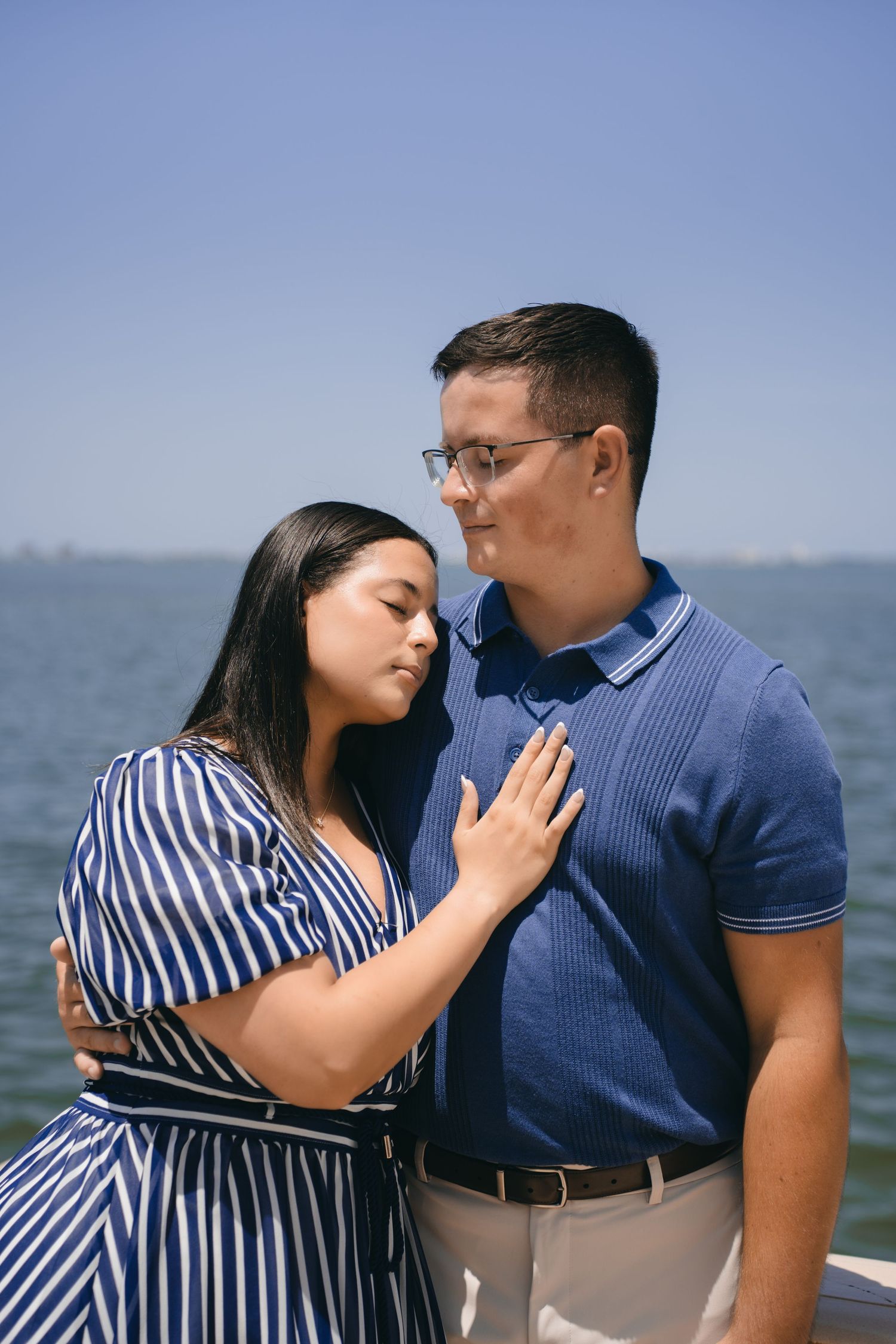 A couple embraces near the water, woman in striped dress rests head on man's chest while he smiles warmly.