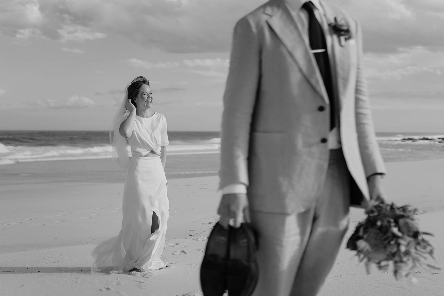 Black and white wedding photo of bride standing on rope swing beside groom in white tuxedo near large tree at dusk.