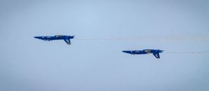 Two Blue Angels jets perform a dramatic crossover maneuver leaving white smoke trails against a pale blue sky.
