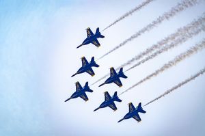 Six Blue Angels jets fly in tight diamond formation, trailing white smoke against a pale blue sky.