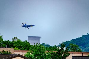 Blue Angels jet flying low over rooftops and trees with a cooling tower visible in the background.