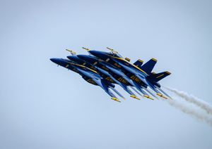 Blue Angels Navy jets flying in tight diamond formation with white smoke trails against a pale blue sky.