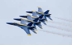Blue Angels jets fly in tight diamond formation leaving white smoke trails against a gray sky.