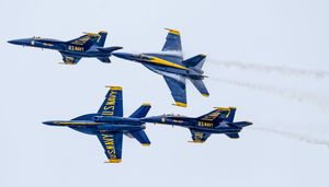 Four U.S. Navy Blue Angels jets fly in close formation, trailing white smoke against a pale sky.