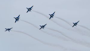 Six Blue Angels aircraft perform a split formation maneuver with crossing smoke trails at an airshow.