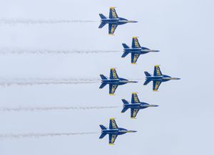Six Blue Angels jets fly in tight diamond formation, leaving white smoke trails against a gray sky.