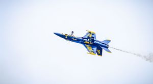 Blue Angels Navy jet performing aerobatic maneuver at airshow, leaving white smoke trail against overcast sky.