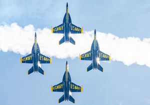 Four US Navy Blue Angels jets fly in diamond formation trailing white smoke against a blue sky.