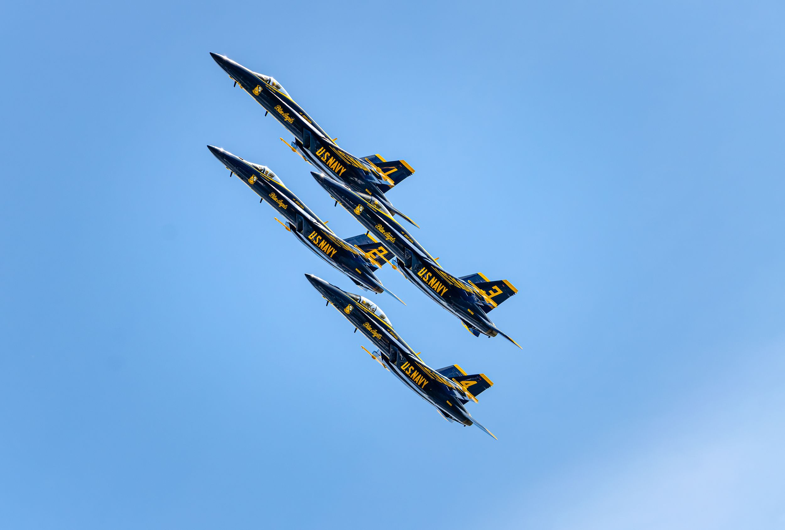 Four Blue Angels Navy jets flying in tight diamond formation against a clear blue sky at an airshow.