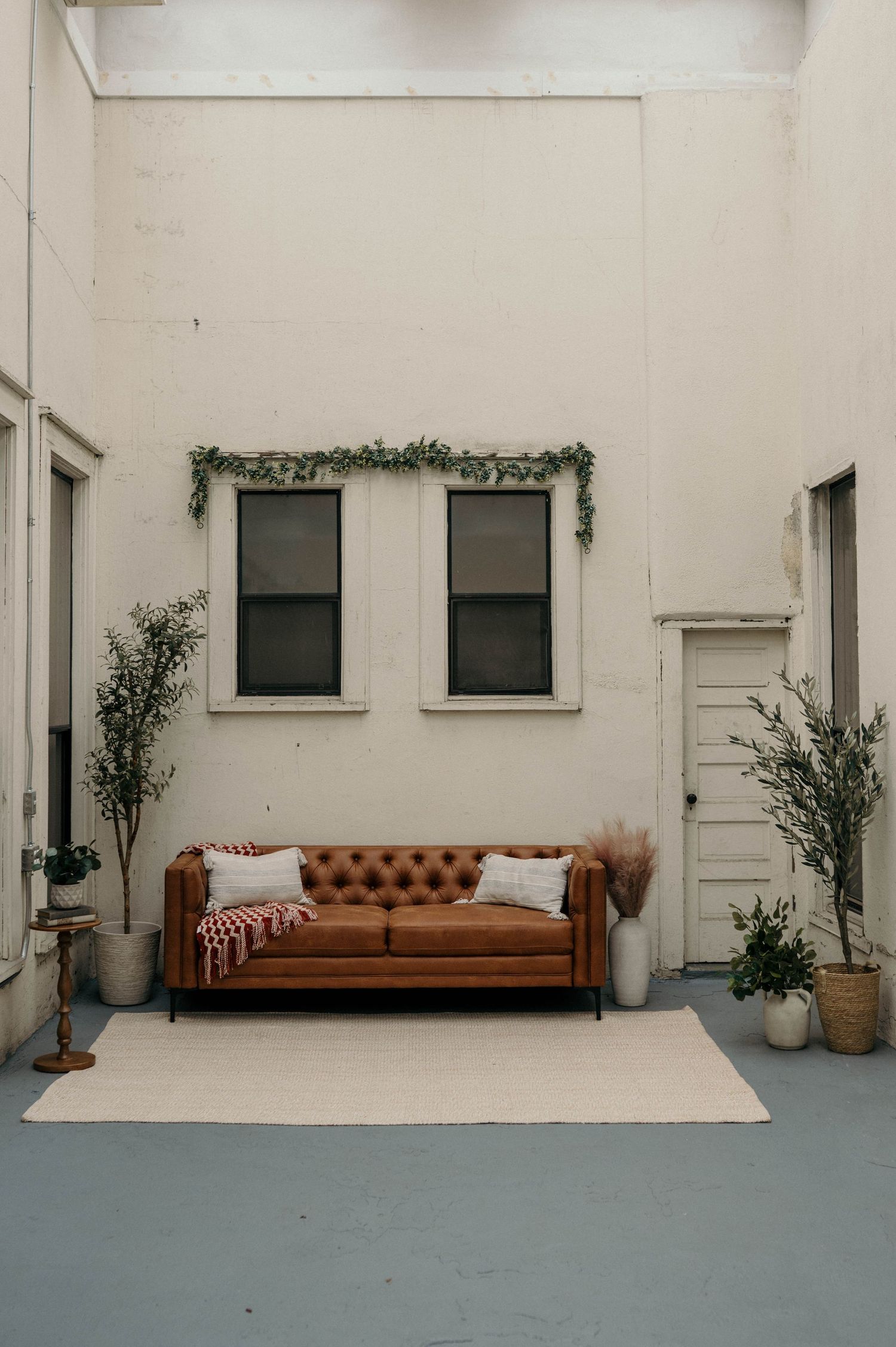 Brown tufted leather Chesterfield sofa on cream rug against white wall with twin windows adorned with green garland.