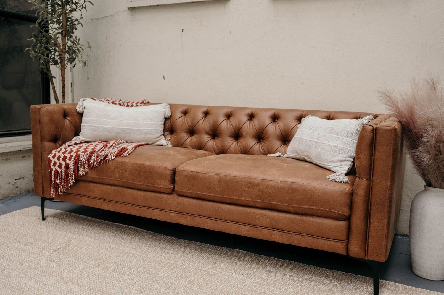 Brown tufted leather Chesterfield sofa with white pillows and red throw blanket on a light area rug.