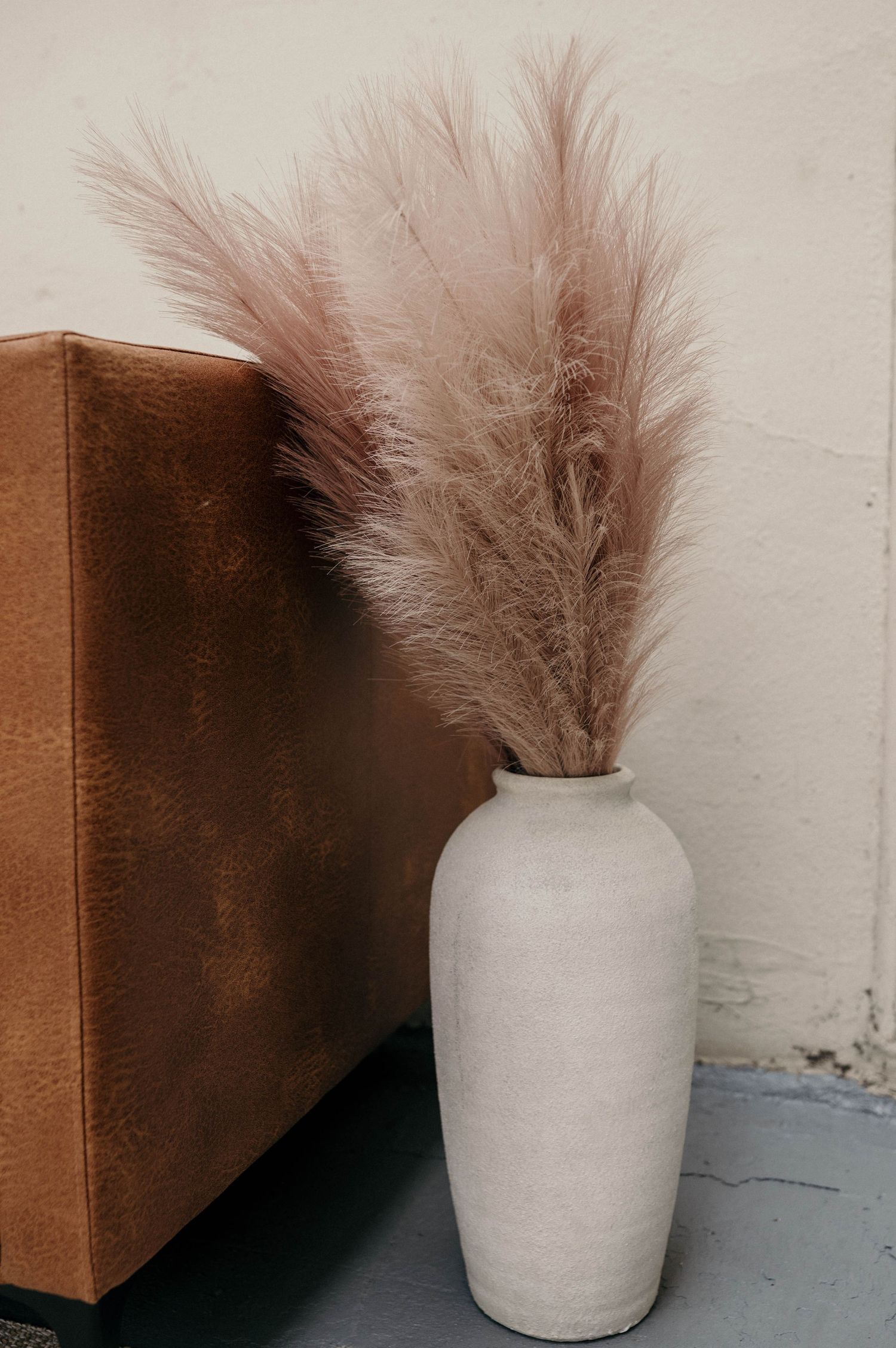 Tall white ceramic vase with fluffy pink pampas grass next to a brown leather object on a gray floor.