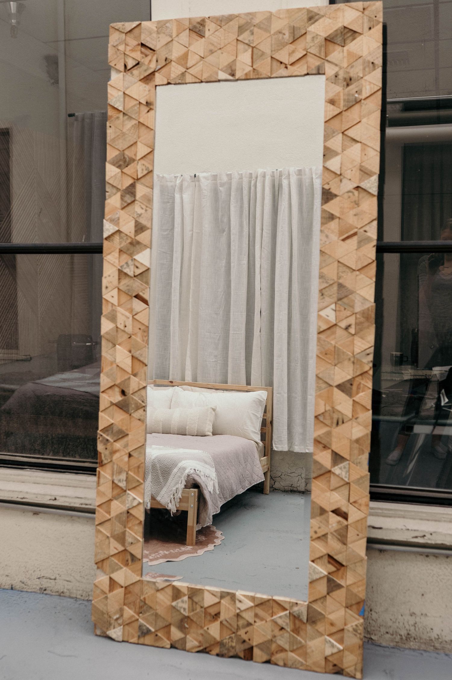 Large floor mirror with geometric wood mosaic frame reflecting a cozy bedroom with white bedding and curtains.